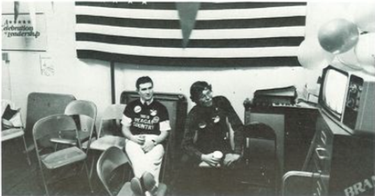 Prompt A. Two students sit in a room full of discarded folding chairs while watching an old tube TV. An American flag hangs on the wall behind them and balloons hover above the TV.